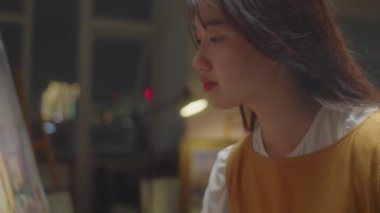 Genç Asyalı kadın ressamın, stüdyoda yeni bir resim üzerinde çalışırken gece şehrinin panoramasını tuvale çizerken yakın çekim.