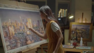 Kadın Asyalı ressamın, yaratıcı stüdyoda resim sehpasında çalışırken gece vakti panoramasını tuvale boyadığı orta boy bir fotoğraf.