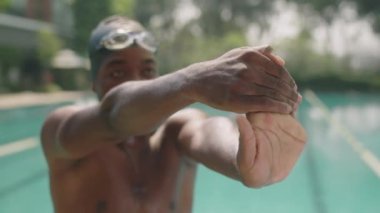 Açık havuzda yüzmeden önce bileklerini ve kollarını açarak ısınan şapkalı ve gözlüklü Afro-Amerikan erkek sporcuyu çek.