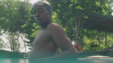 Açık havuzun kenarında kameraya poz verirken şapkalı ve gözlüklü profesyonel Afro-Amerikan yüzücünün düşük açılı görüntüsü.