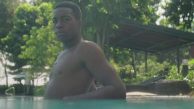 Orta boy Afrika kökenli Amerikalı bir adam yüzme havuzunun kenarına yaslanmış ve yaz günü dışarıda kameraya poz veriyor.