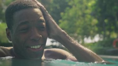 Genç ve neşeli Afro-Amerikalı adamın yüzme havuzunda serinlerken ve yaz gününü dışarıda geçirirken mutlu bir şekilde gülümserken yakın plan çekimleri.