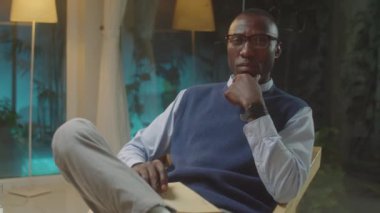 Siyah erkek psikoloğun akşam saatlerinde ofisinde oturup kameraya özgüvenle baktığı ve çenesinin altındaki elle poz verdiği orta ölçekli bir fotoğraf.