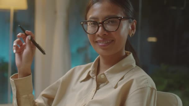 グラスにアジアの女性セラピストのチェストアップポートレートショットとペンを保持し 夜中に彼女のオフィスで働いている間笑顔でカメラのためにポーズするエレガントな衣装 — ストック動画