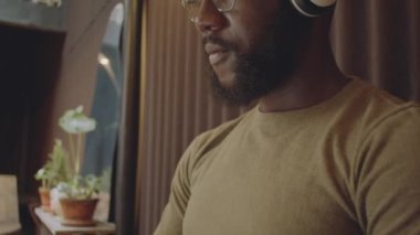 Genç Afrikalı Amerikalı bir adamın kablosuz kulaklıkla müzik dinlerken ve kafede otururken dizüstü bilgisayarda çalışırken orta boy yakın çekimi.