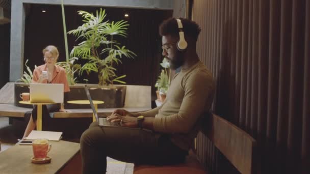 放大侧视拍摄的年轻的非洲裔美国人坐在咖啡店的无线耳机里 一边在笔记本电脑上打字一边远程工作 — 图库视频影像