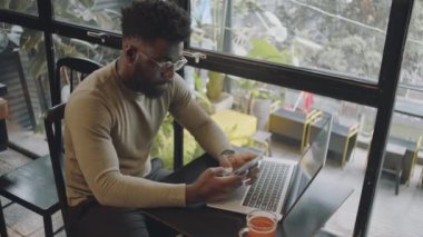 Genç Afro-Amerikan iş adamının günlük kıyafetleri ve gözlükleriyle akıllı telefondan mesajlaşırken kafeteryada dizüstü bilgisayar ve çay fincanı ile otururken yüksek açılı görüntüsü.