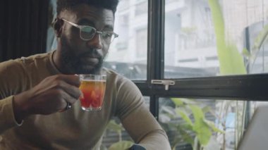 Genç Afro-Amerikan iş adamının günlük kıyafetleri ve gözlükleriyle çay içerken ve kafede uzaktan çalışırken dizüstü bilgisayar ekranına bakarken orta boy yakın çekimi.