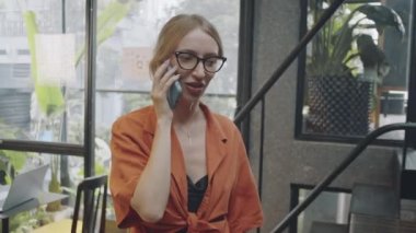 Modern çalışma alanlarında yürürken ve cep telefonuyla konuşurken gözlüklü genç bir kadının orta boy fotoğrafı.