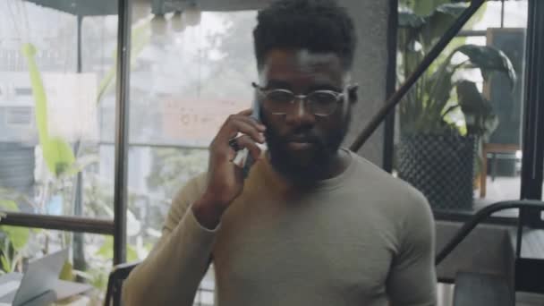 コワーキングスペースを歩き 携帯電話で話すカジュアルウェアと眼鏡を着た若いアフリカ系アメリカ人のメディアショット — ストック動画