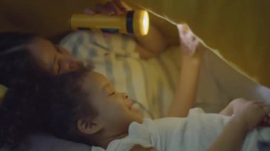 Annesiyle birlikte geceleri battaniyenin altında el feneri tutarken gülümseyen ve oyun oynayan Asyalı küçük kızın orta boy yakın çekimi.