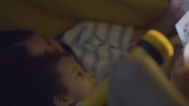 アジアの母親と小さな娘を愛し 暗い部屋でベッドに横たわり 寝る前に懐中電灯と読書本を保持する中間クローズアップショット — ストック動画