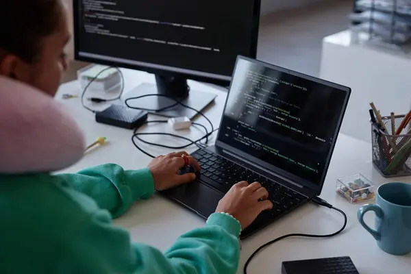 Programmer sitting at her workplace with computers and writing codes on laptop in office