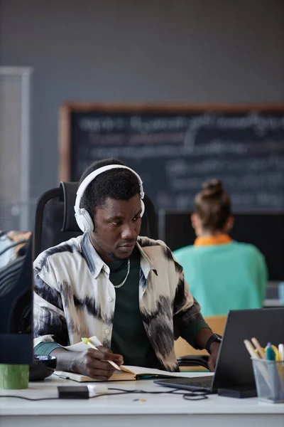 Vertical image of African American programmer making notes while working on laptop at his workplace