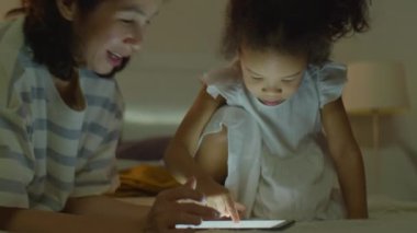 Gülümseyen Asyalı anne ve sevimli küçük kız akşam evde dijital tablette birlikte oynuyorlar.