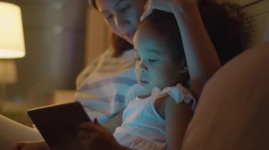 Asyalı annenin küçük kızına sarıldığı, kafasını okşadığı ve akıllı telefonu kız gibi evde yatağında dijital tablette çizgi film izlerken çektiği orta boy yakın çekim.