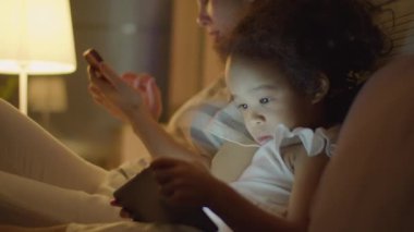 Annesi evde yatağında telefon kullanırken Asyalı küçük kızın dijital tabletle çizgi film izlediği orta boy yakın çekim.