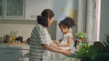 Neşeli Asyalı kadın lavaboda bulaşıkları yıkıyor ve mutfak tezgahında oturan ve tabakları havluyla kurutan tatlı küçük kızıyla konuşuyor.