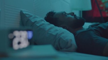 Genç adamın karanlık bir odada yatarken ve gece uykusuzluk çekerken dijital saate bakarken çekilen fotoğraf.