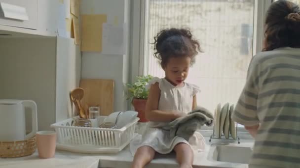 可爱的亚洲幼儿女孩坐在厨房的台面上 用毛巾擦干盘子 同时帮助妈妈在家里洗碗 — 图库视频影像