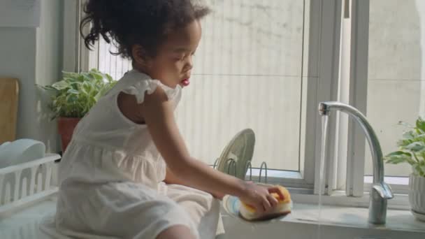 アジアの母親のハンドヘルドカメラショットは シンクで座っているかわいい小さな娘のようにきれいな水でプレートを洗い流し 梯子と遊び 自宅でキッチンで料理をするのを助けます — ストック動画