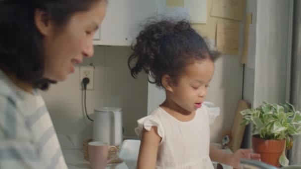 キッチンカウンターに座って座って 母親が料理をしている間に梯子と水で遊ぶ愛らしい小さなアジアの少女 — ストック動画