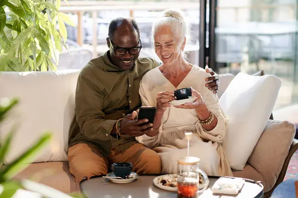 Happy senior couple drinking coffee in cafe and having video call on smartphone