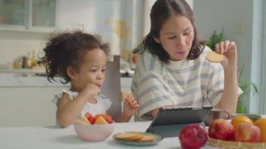 Orta boy Asyalı kız ve anne mutfak masasında birlikte otururken, kurabiye yerken, çay içerken ve dijital tablette bir şeyler izlerken.