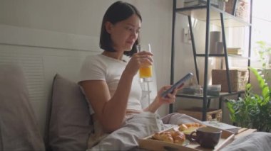 Genç bir kadın yatakta kahvaltı yapıyor, portakal suyu içiyor ve sabah evde cep telefonu kullanıyor.