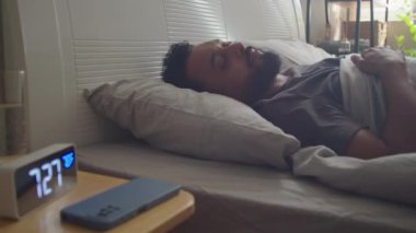 Genç sakallı bir adam sabah yatağında uyanır, çalar saati kapatır ve evde akıllı telefon kullanır.