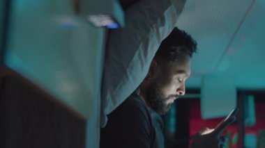 Yatakta telefonla mesajlaşan, sonra battaniyenin altında uyuyan genç sakallı adamın dikey görüntüsü.