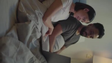 Genç bir çiftin dizüstü bilgisayardan film izlerken akşamları yatakta otururken çekilmiş dikey görüntüleri.