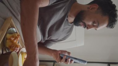 Yatakta kahvaltı yapan, portakal suyu içen ve evde akıllı telefondan haber okuyan bir adamın dikey görüntüsü.