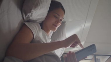 Yatakta yatan genç bir kadının dikey görüntüsü ve sabah uyandıktan sonra sosyal medyada akıllı telefondan kaydırma.
