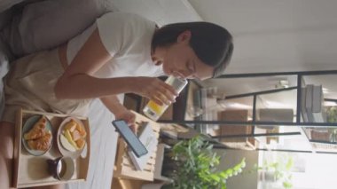Genç bir kadının yatakta kahvaltı ederken, portakal suyu içerken, meyve yerken ve sabahları evde akıllı telefonun dokunmatik ekranında kayarken dikey yüksek açılı görüntüsü.