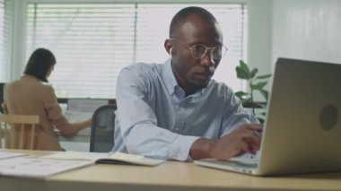 Resmi gömlek, gözlük ve kablosuz kulaklıkla ofisteki laptopta çalışan Afrikalı Amerikalı iş adamının orta boy fotoğrafı.