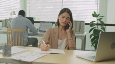 Ofiste çalışırken cep telefonuyla konuşan ve notlar alan genç bayan yöneticinin orta boy fotoğrafı.
