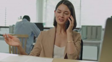 Genç zarif iş kadınının ofis masasında oturup iş günü cep telefonuyla konuştuğu orta boy bir fotoğraf.