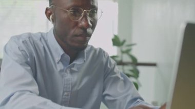 Resmi gömlekli, gözlüklü ve kulaklıklı Afro-Amerikan iş adamının laptopta daktilo ederken, düşünceli bir şekilde ekrana bakarken ve iş günü çenesini sıvazlarken görüntüsünü kaldır.