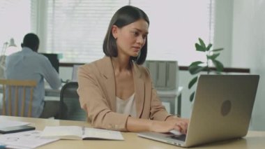 Ofiste dizüstü bilgisayarda çalışan genç ve güzel bir iş kadınının orta ölçekli bir resmi, sonra kamera önünde poz vermek ve gülümsemek.