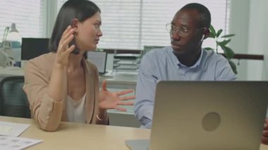 Kafkasyalı genç iş kadınının ofis masasında yan yana otururken Afrikalı Amerikalı iş arkadaşına dizüstü bilgisayardaki projeyi açıklarken orta boy fotoğrafı.