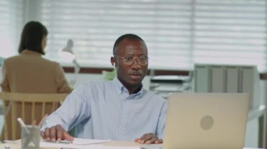 Resmi giyimli, gözlüklü ve kablosuz kulaklıklı Afrikalı Amerikalı işadamının ofiste çalışırken bilgisayardan yaptığı görüntüler.