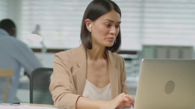 Formalite ve kablosuz kulaklık takmış genç bir iş kadınının iş günlerinde laptoptan internet üzerinden konuşma görüntüsü.