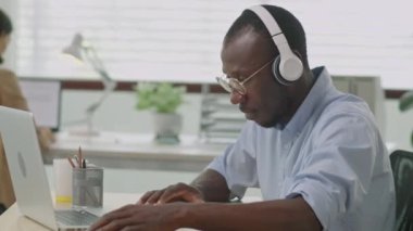Kulaklık takmış, müzik dinleyen, dizüstü bilgisayarda çalışan ve ofis masasındaki belgeleri kontrol eden Afrikalı Amerikalı iş adamının orta boy fotoğrafı.