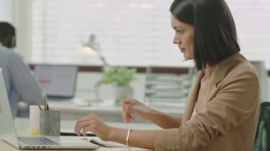 Genç bir iş kadınının dizüstü bilgisayarında yazı yazarken ve ofiste çalışırken notlar alırken orta ölçekli bir yan görüntüsü.