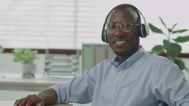 身着无线耳机坐在办公室里 摆出一副拍照和微笑的样子的快乐的非洲裔美国商人的中肖像画 — 图库视频影像