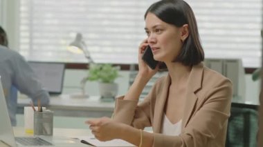 Ofiste çalışan ve cep telefonuyla konuşan genç profesyonel iş kadınının orta boy fotoğrafı.