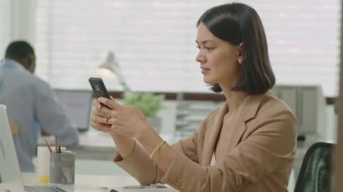 Güzel bir iş kadınının ofis masasında oturduğu, cep telefonuna baktığı ya da iş günü mesajlaştığı orta ölçekli bir yan görüntü.