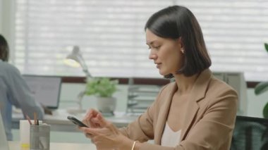 Genç profesyonel iş kadınının akıllı telefondan mesajlaşmasını, sonra kameraya poz vermesini ve iş yerinde otururken gülümsemesini.