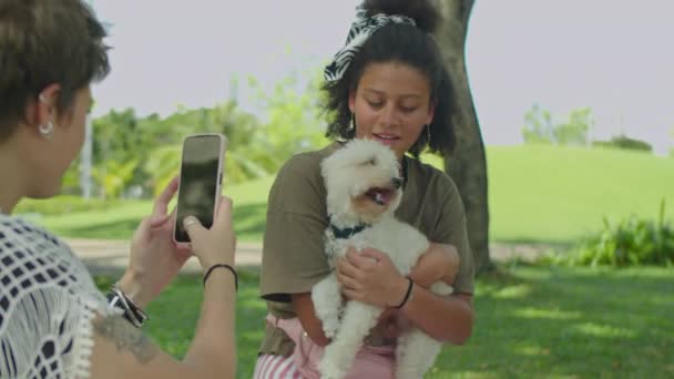 女孩和可爱的狗摆姿势拍照 然后在公园野餐的时候和朋友们用智能手机讨论照片 — 图库视频影像